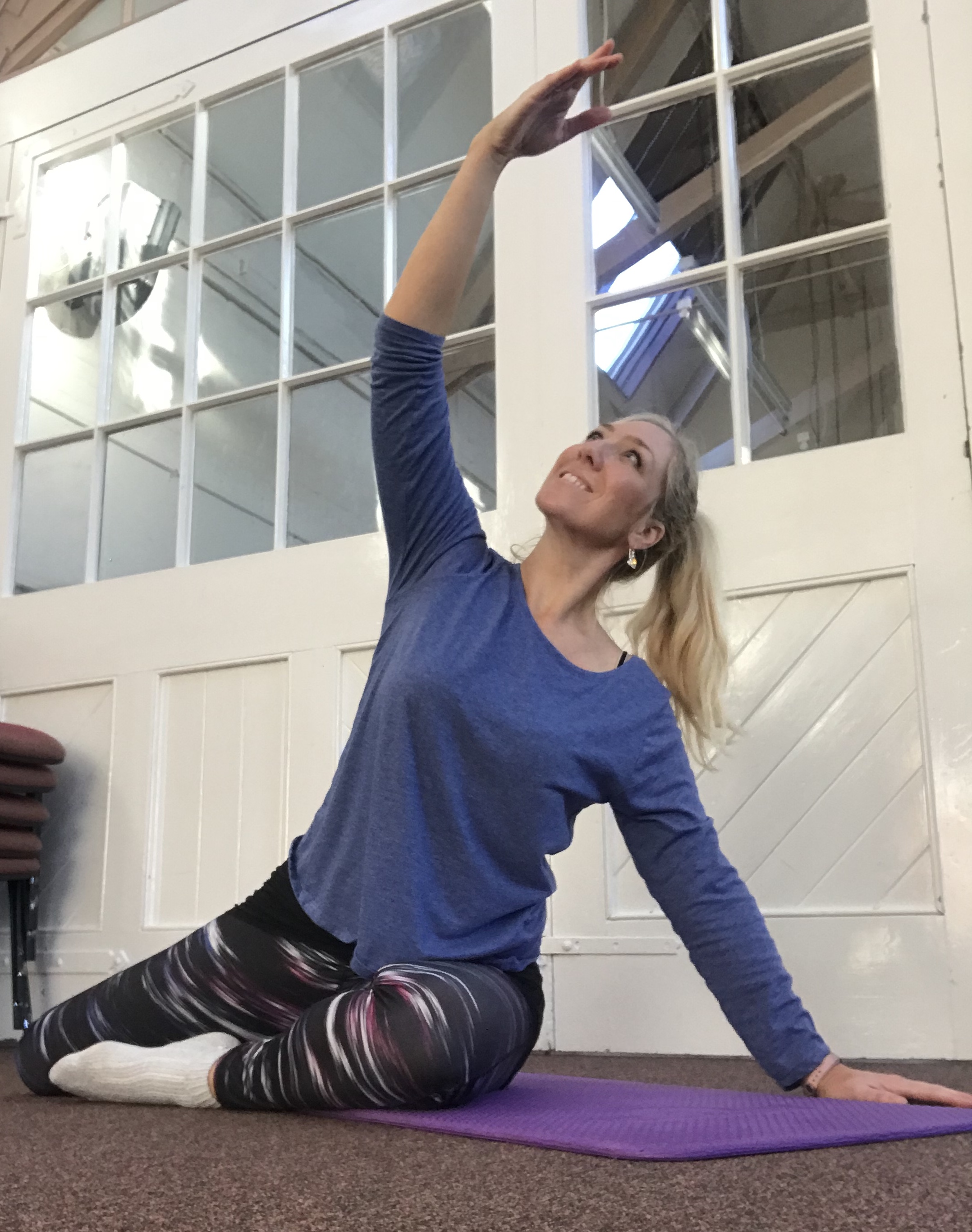 Cheryl Bromley in a kneeling side-bend stretch on a mat, in a hall with tall white windows and a wooden floor