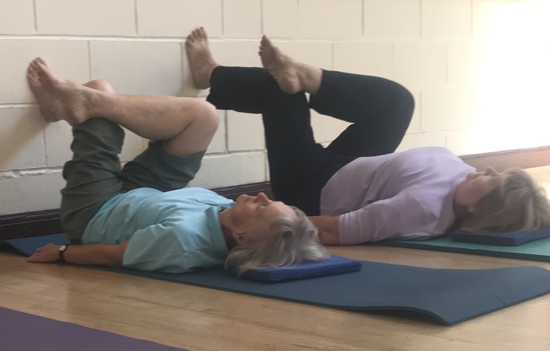 Two clients side by side on mats against a painted brick wall, legs raised in tabletop position