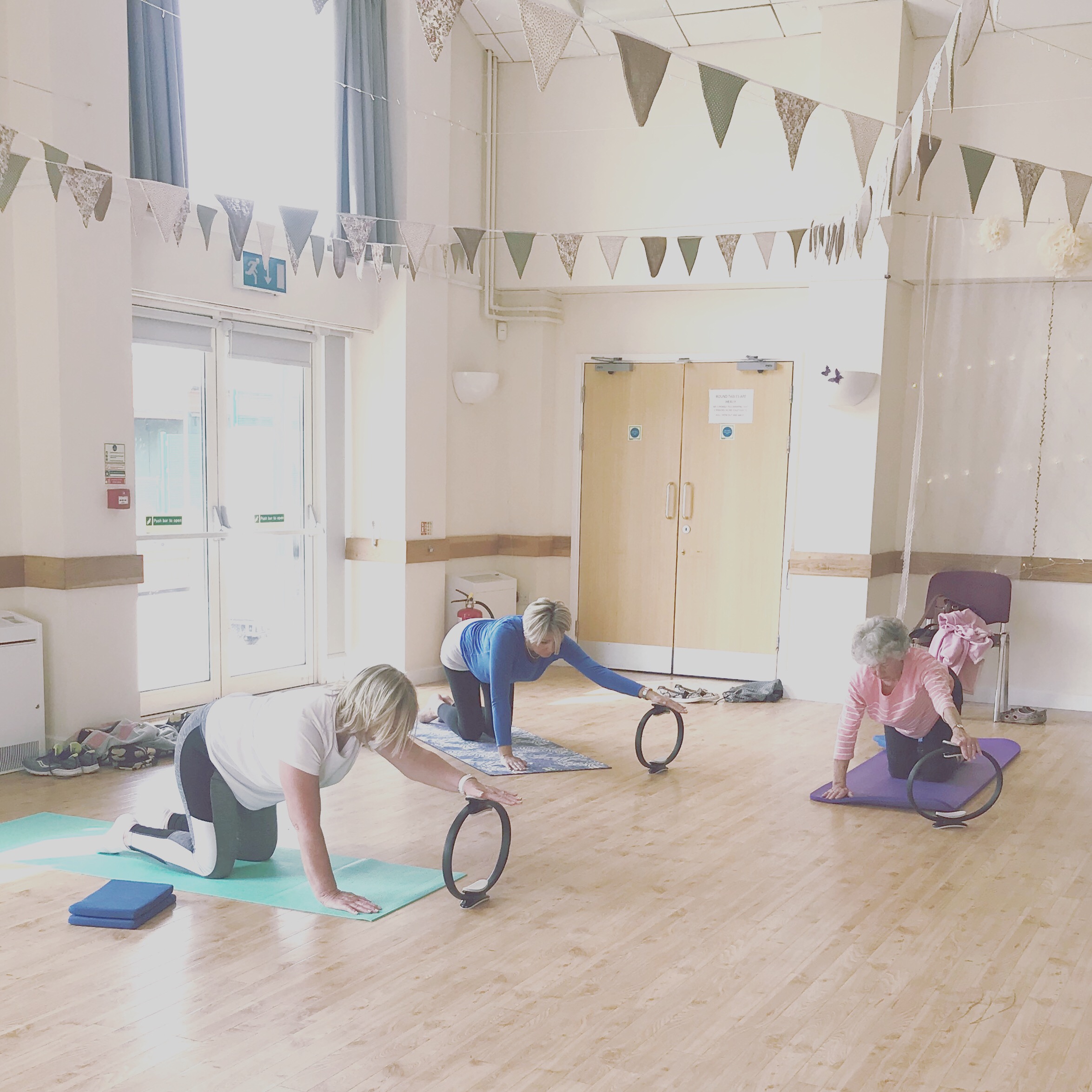 Three clients on mats holding Pilates rings in a village hall with bunting strung across the ceiling